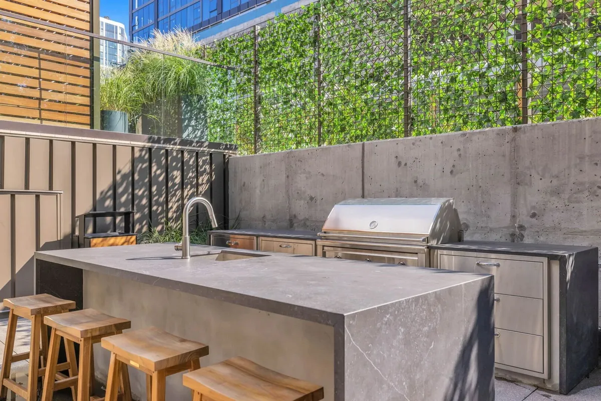 An outdoor kitchen island equipped with a stainless steel grill, wooden stools, and a backdrop of greenery. The space is set for barbecuing, dining, and enjoying the outdoors with friends or neighbors.