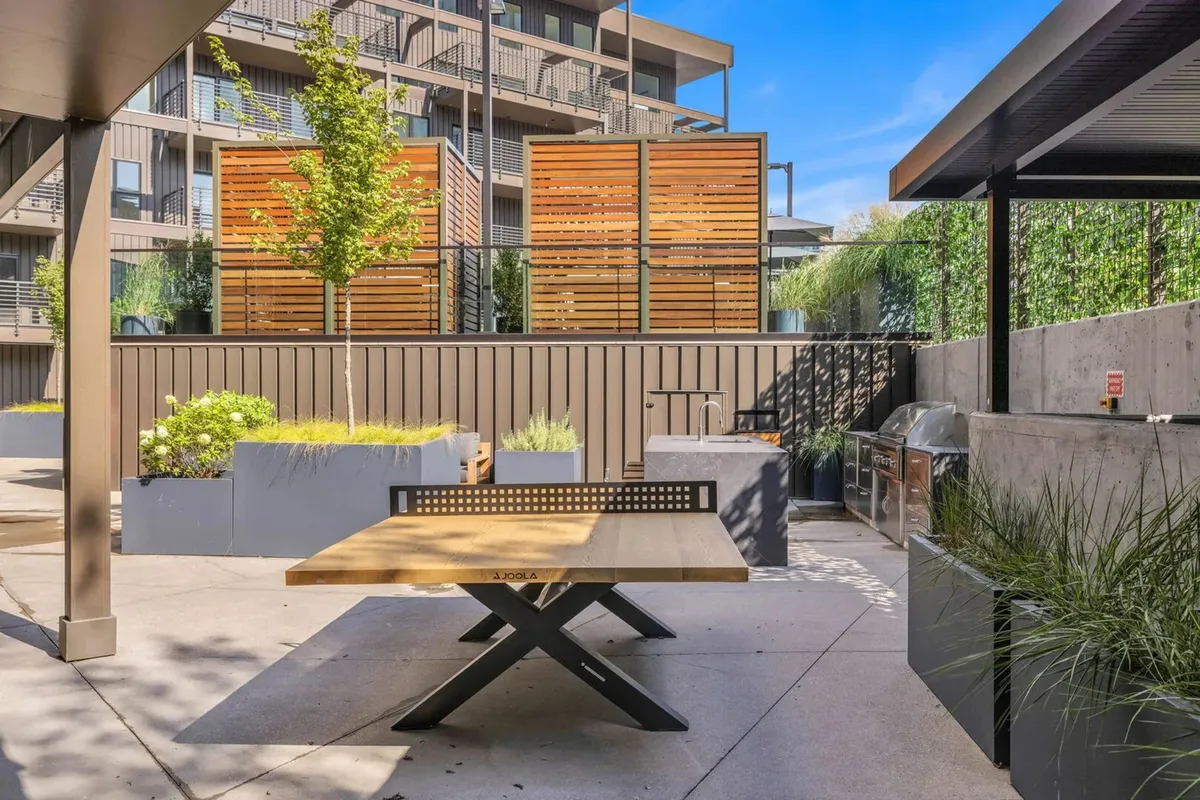 An inviting exterior scene of a balcony dining area featuring a wooden table, green plants, wooden screens, and a grill area. The setting is ideal for dining outdoors, surrounded by greenery and fresh air.