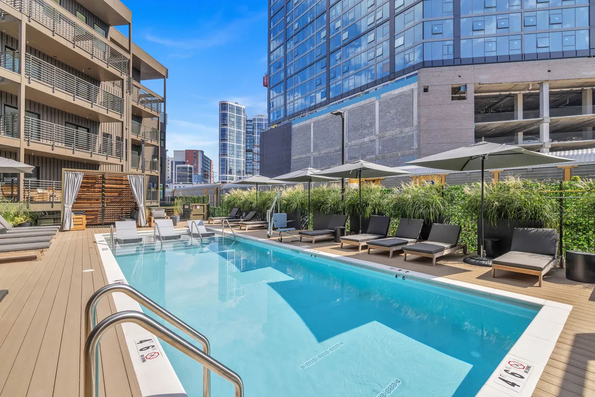 A sophisticated exterior pool area featuring modern lounge chairs, umbrellas, lush greenery, and impressive building architecture. The scene is relaxed but vibrant, suggesting a place for both socializing and unwinding after work.