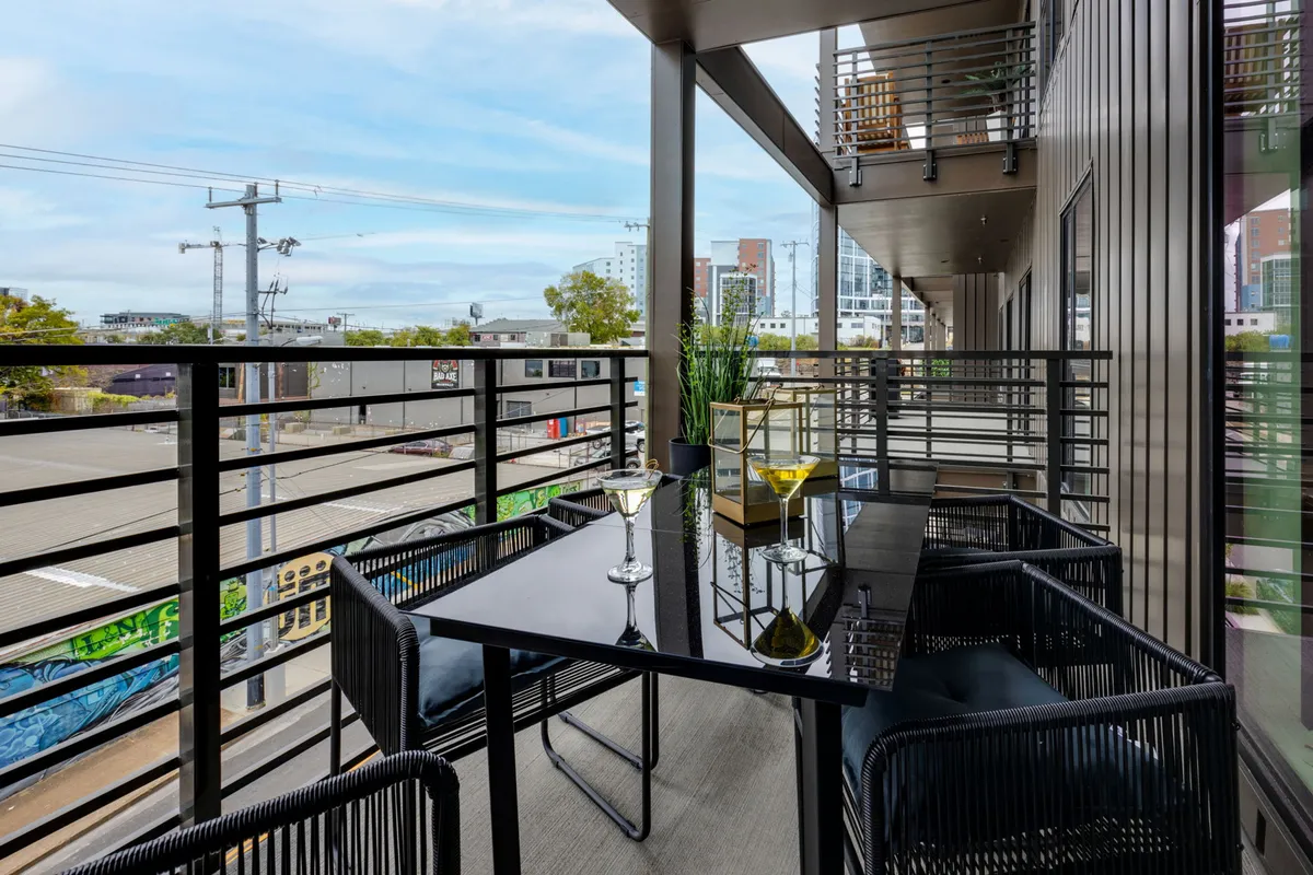A modern balcony set with a glass table and drinks, contemporary chairs, and an urban view of the city skyline. The mood is relaxed and sophisticated, ideal for enjoying sunset cocktails or morning coffee above the city bustle.