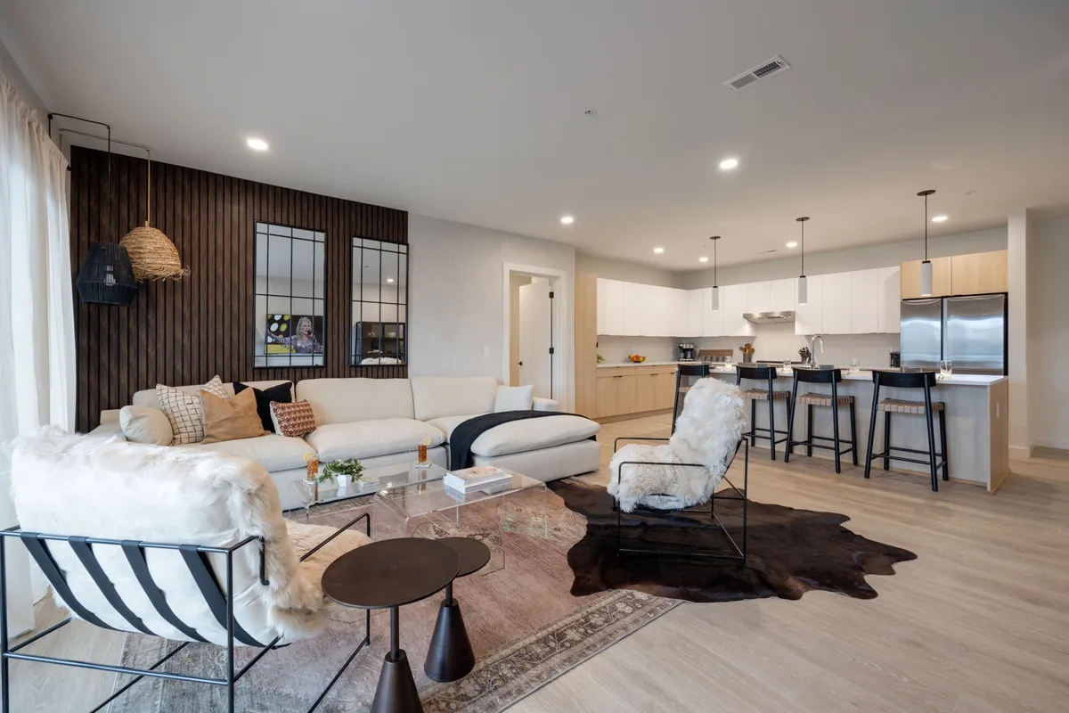 The image showcases a spacious, open-concept living area with a large, inviting sectional sofa accented by vibrant decorative pillows. A striking wooden accent wall adds warmth and Nashville character, while the cowhide rug infuses a touch of Southern flair. Bar stools at the stylish kitchen island suggest a seamless space for socializing, dining, or relaxing, ideal for group gatherings.