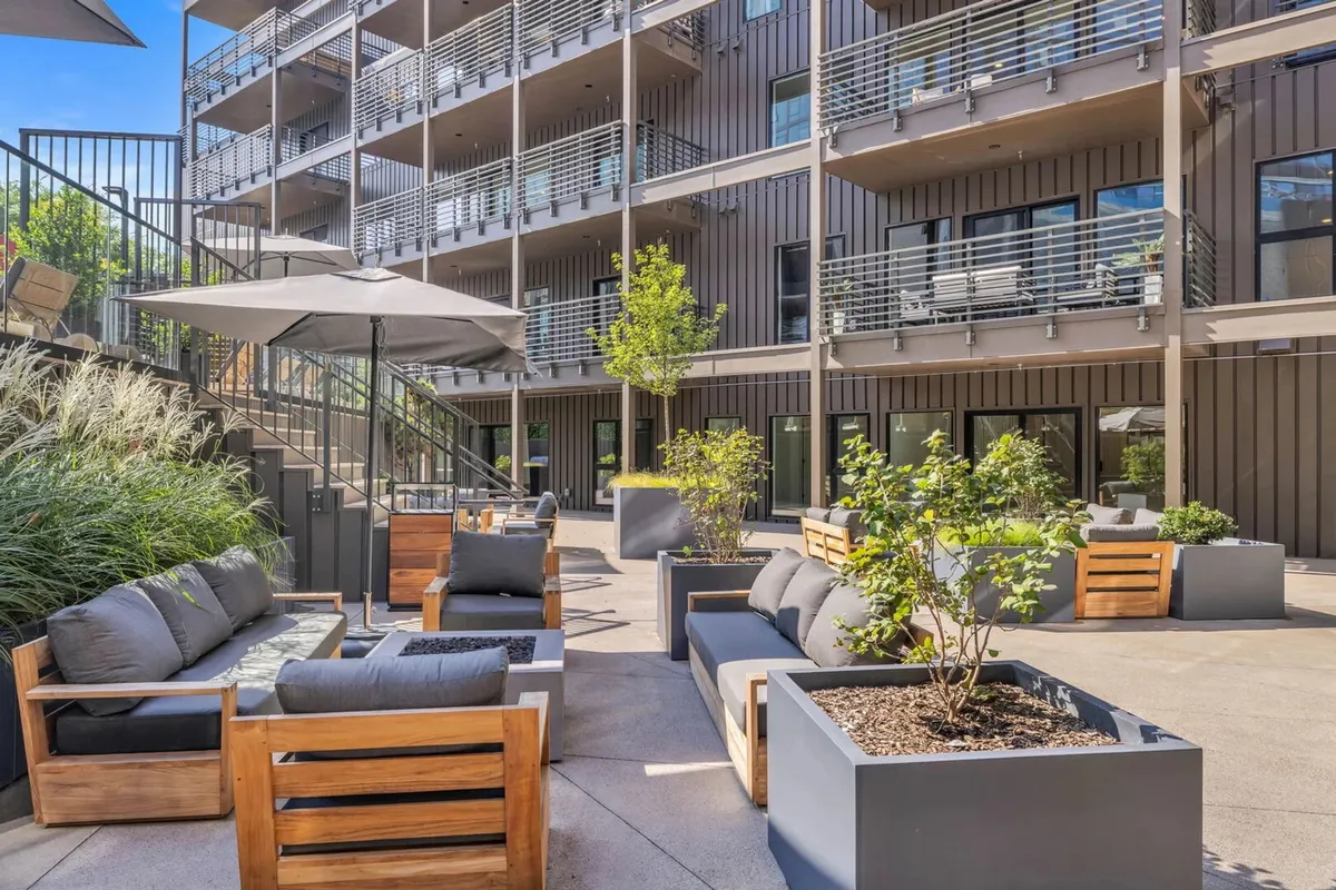 The image features a modern outdoor seating area with comfortable arrangements, decorative planters, and a staircase leading to upper levels. The space is designed for group gatherings, relaxation, and conversations under the open sky.