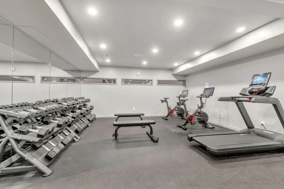 A stylish, modern gym interior featuring a dumbbell rack, treadmills, stationary bikes, and a weight bench. The space feels motivational and ready for both solo workouts and group fitness sessions.