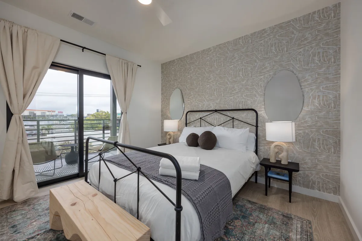 A serene bedroom featuring a plush bed with decorative pillows, large windows allowing abundant natural light, a textured accent wall, and nightstands with lamps. The space exudes calm and comfort, providing a welcoming atmosphere for rest.