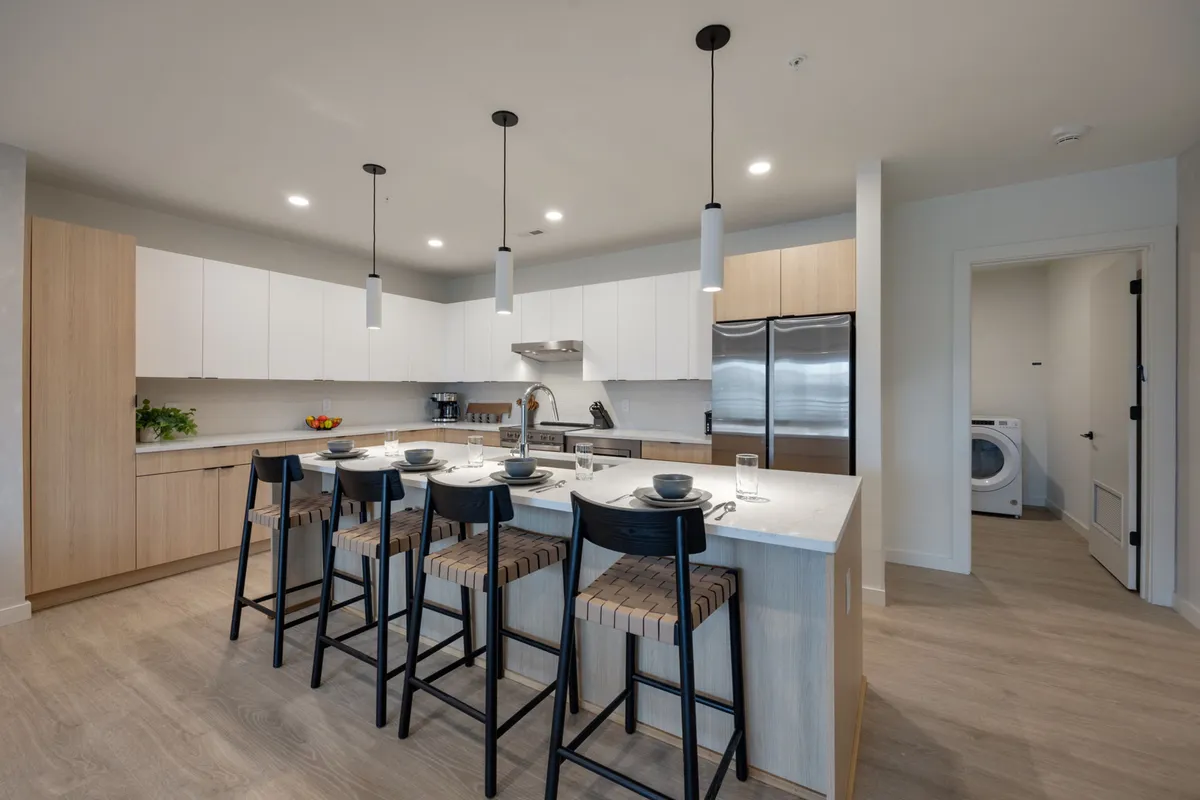 A bright, modern kitchen featuring a central island with bar stools, stainless steel appliances, and a welcoming dining setup. The space is designed for both cooking and socializing, with ample seating and plenty of natural light.
