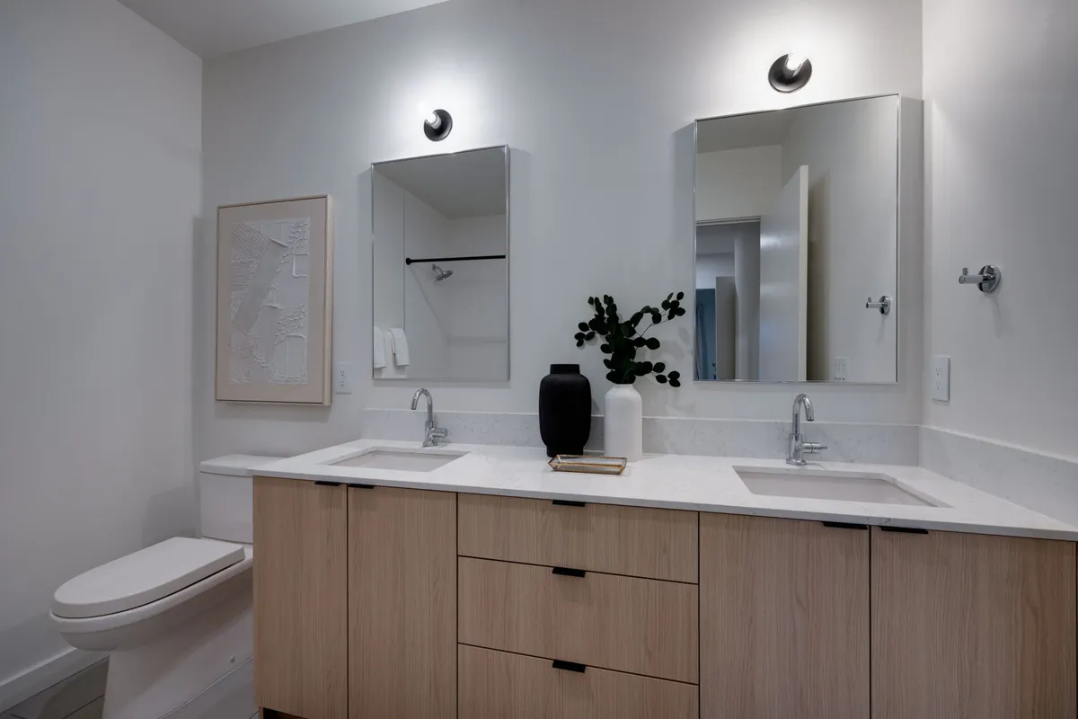 A modern bathroom with a double sink vanity, large mirrors, a decorative plant, and framed artwork. The setting is calm and spa-like, perfect for efficient morning routines or relaxing self-care.