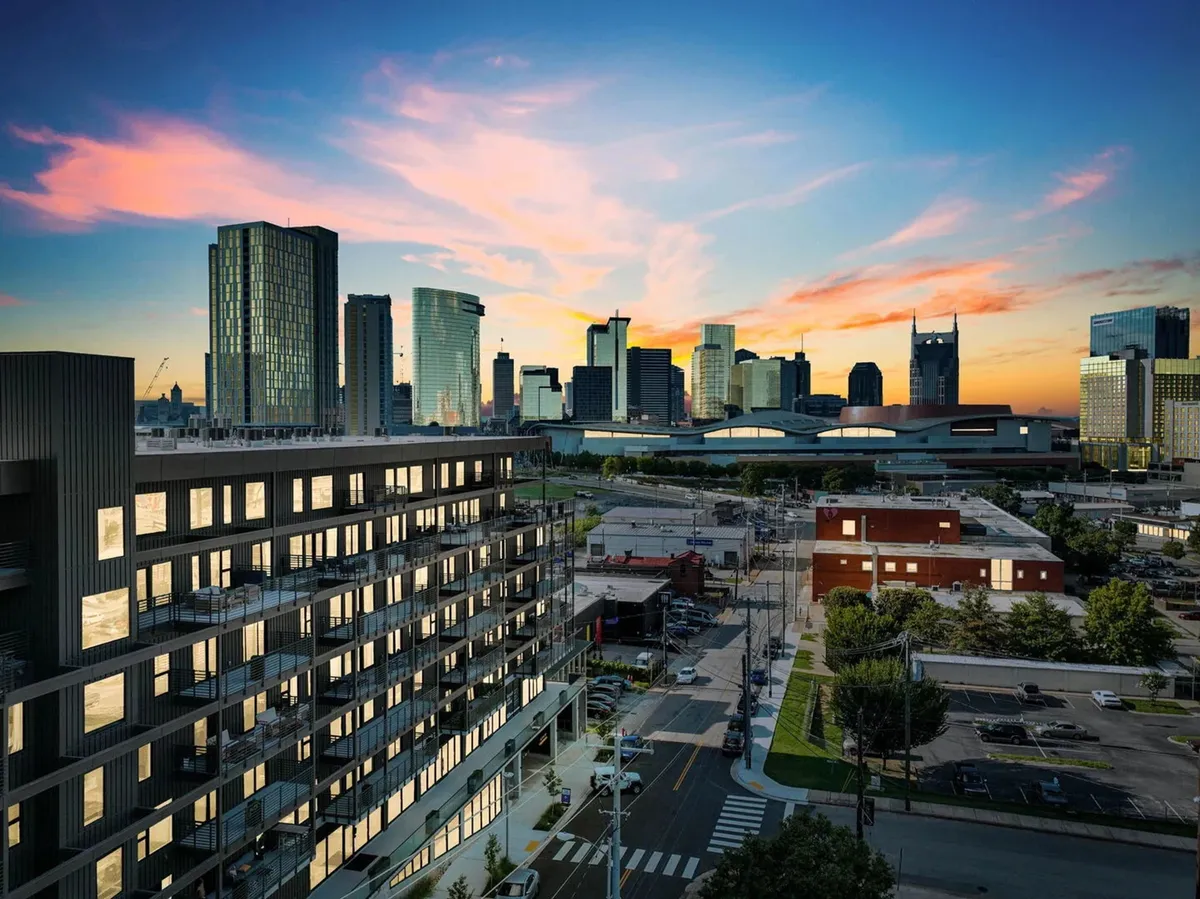 A modern apartment building set against a vibrant city skyline at sunset, with balconies overlooking the bustling urban landscape. The sky is painted in warm dusk hues, and the energy of the city is palpable, hinting at nearby restaurants, nightlife, and exploration opportunities.